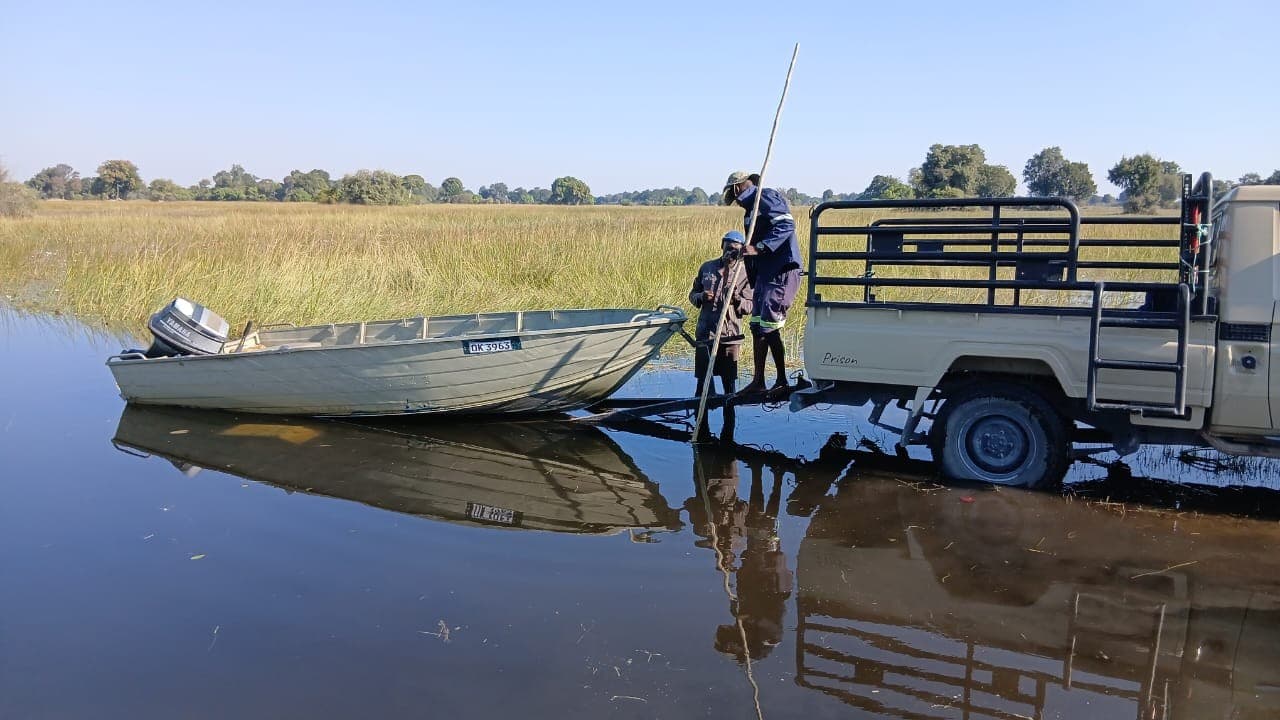 Boat meeting a 4x4 vehicle at the water's edge for a guest transfer at Samonyana, with still water reflecting the scene.