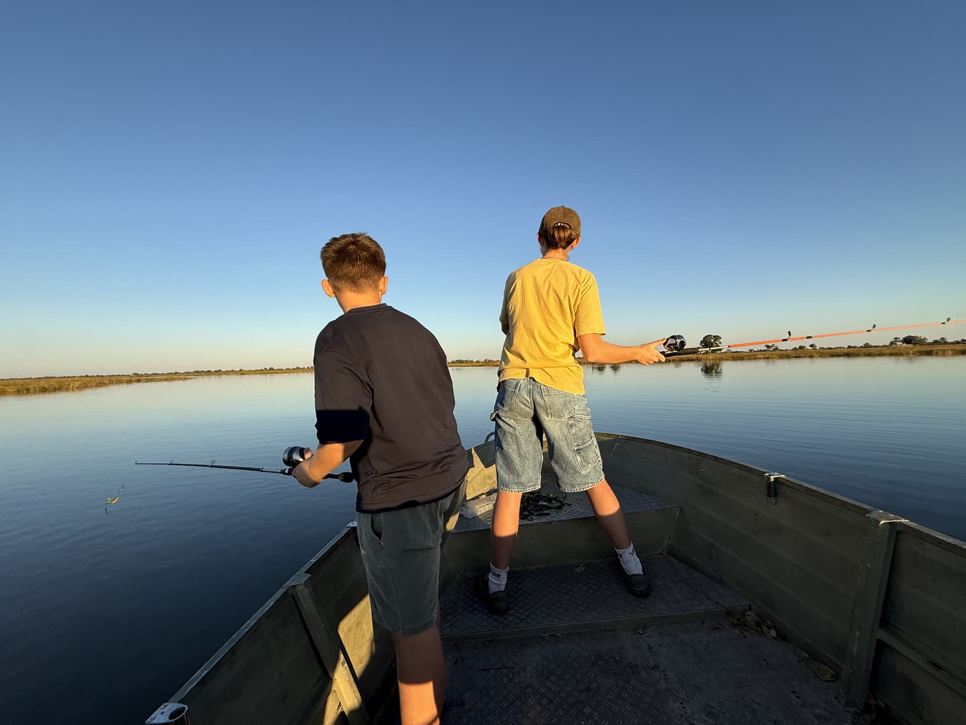 Two young guests fishing from a boat on a calm Okavango Delta channel at golden hour.