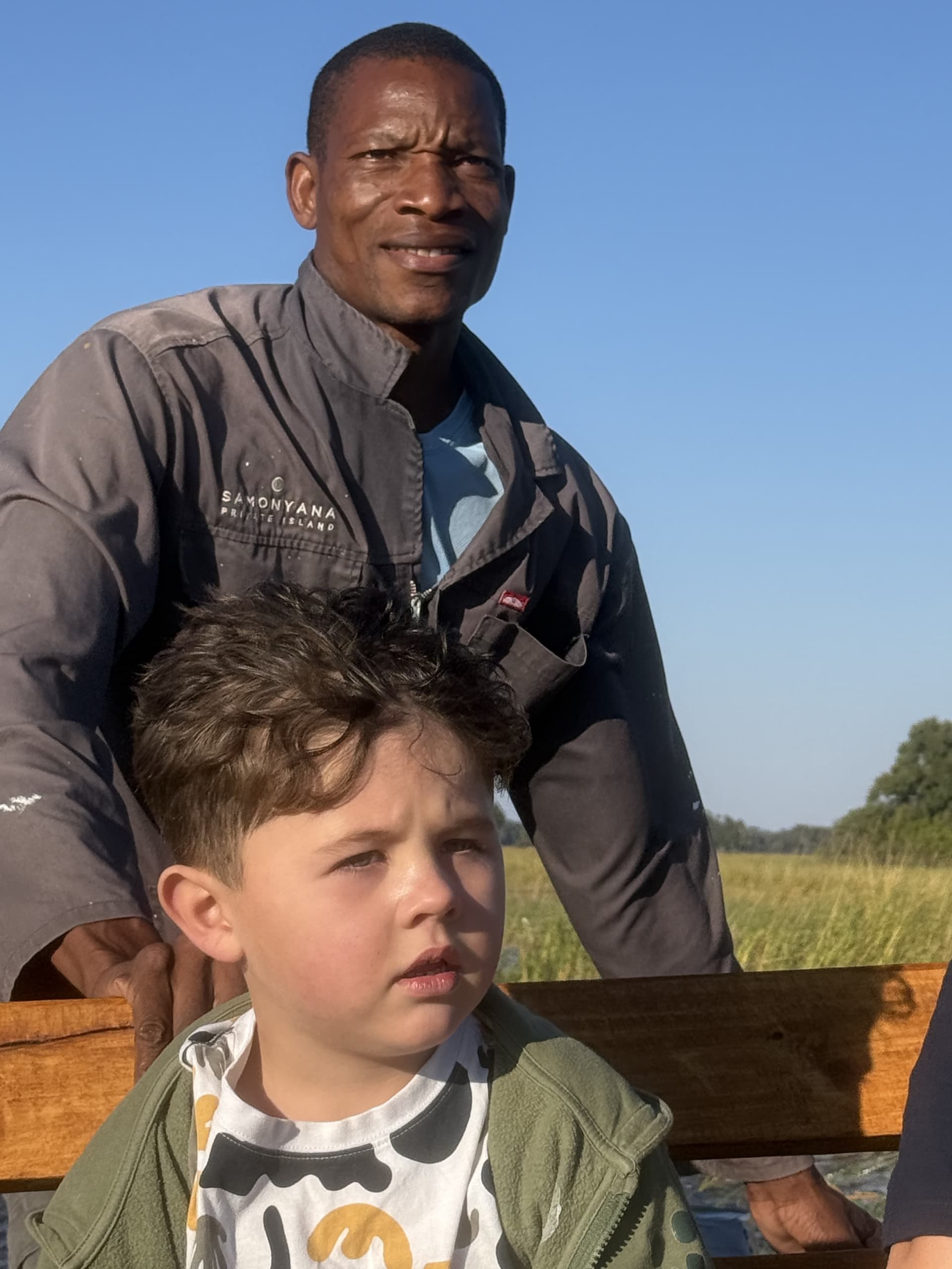 Safari guide standing behind a young guest on an open-vehicle game drive under clear skies at Samonyana Private Island.