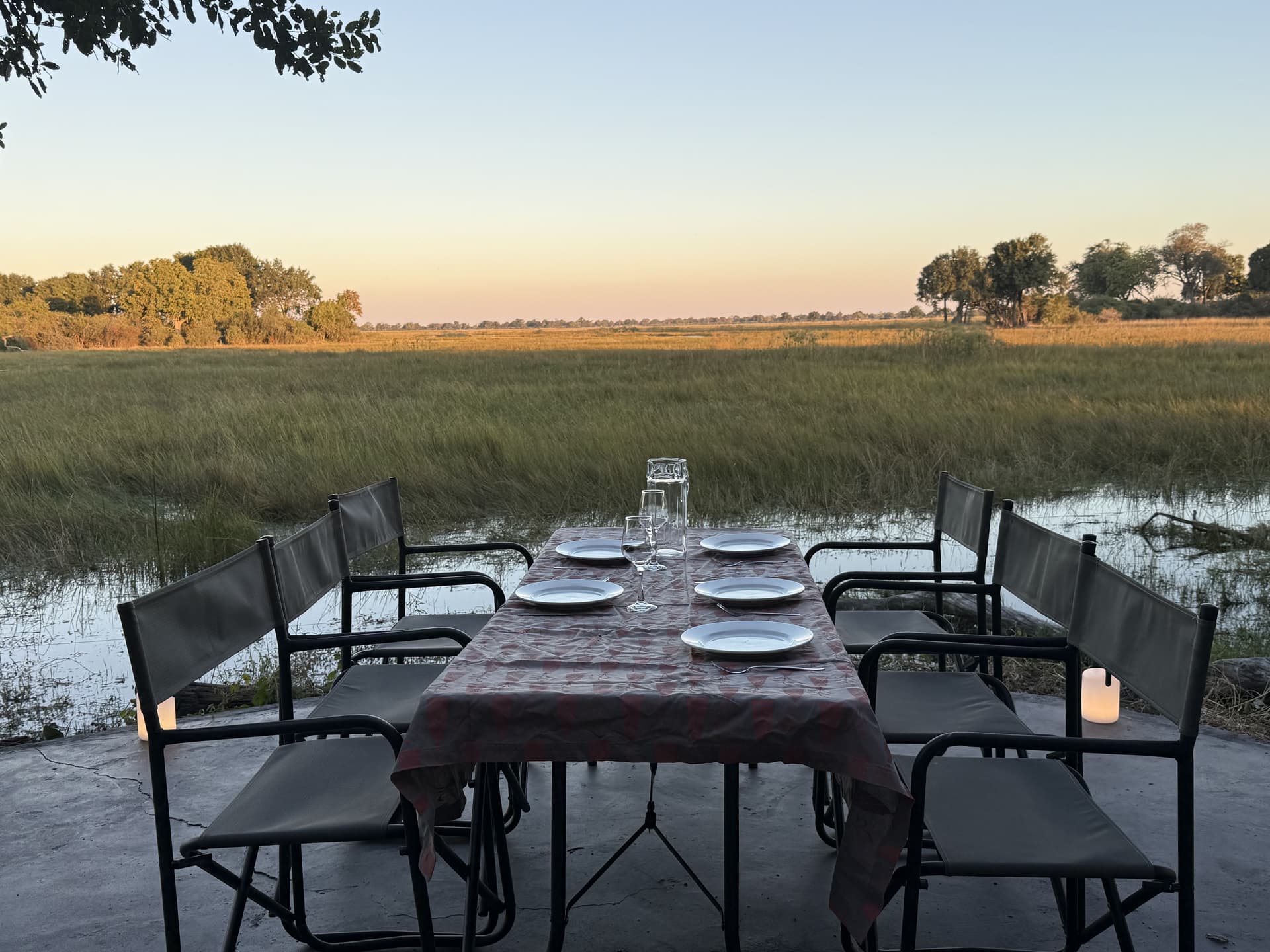 Open-air dining table set with candles on a circular deck overlooking the Okavango Delta floodplain at sunset